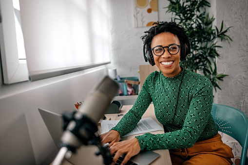 Portrait of smiling young women typing on laptop and recording podcast while speaking in microphone at home
